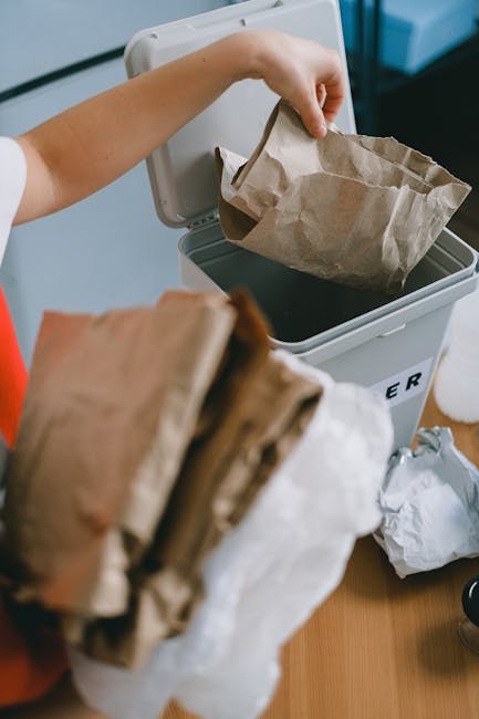 A person disposing of paper waste into a grey rubbish bin with a partially open lid, which is situated on a wooden surface indoors. The individual’s arm and hand are visible, holding a crumpled brow