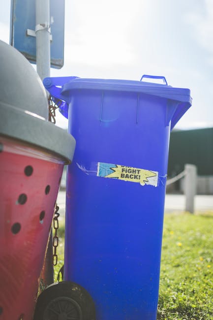 A person disposing of paper waste into a grey rubbish bin with a partially open lid, which is situated on a wooden surface indoors. The individual’s arm and hand are visible, holding a crumpled brow